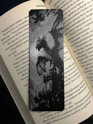 Zakładka 3D z czarno-szarym smokiem na tle księżyca i gotyckiego zamku, włożona w książkę